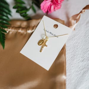 May include: A gold-colored necklace with a cross and a religious medallion charm, displayed on a white card. The card is placed on a gold satin pouch, with pink flowers and green leaves in the background. The text "Catholic Made Goods" is visible on the card.