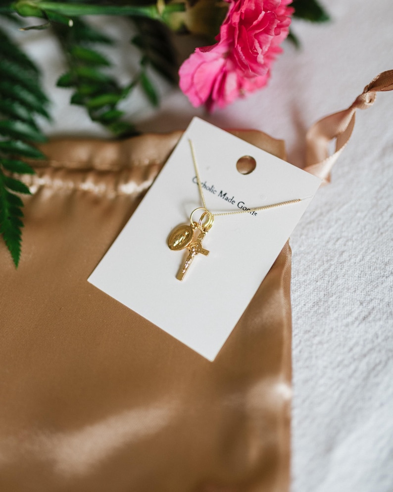 May include: A gold-colored necklace with a cross and a religious medallion charm, displayed on a white card. The card is placed on a gold satin pouch, with pink flowers and green leaves in the background. The text "Catholic Made Goods" is visible on the card.