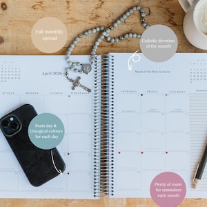 May include: Open spiral-bound planner for April 2026, featuring a full monthly spread, feast day and liturgical colors, and room for reminders. A rosary and a smartphone rest on the pages, with a mug of coffee nearby.