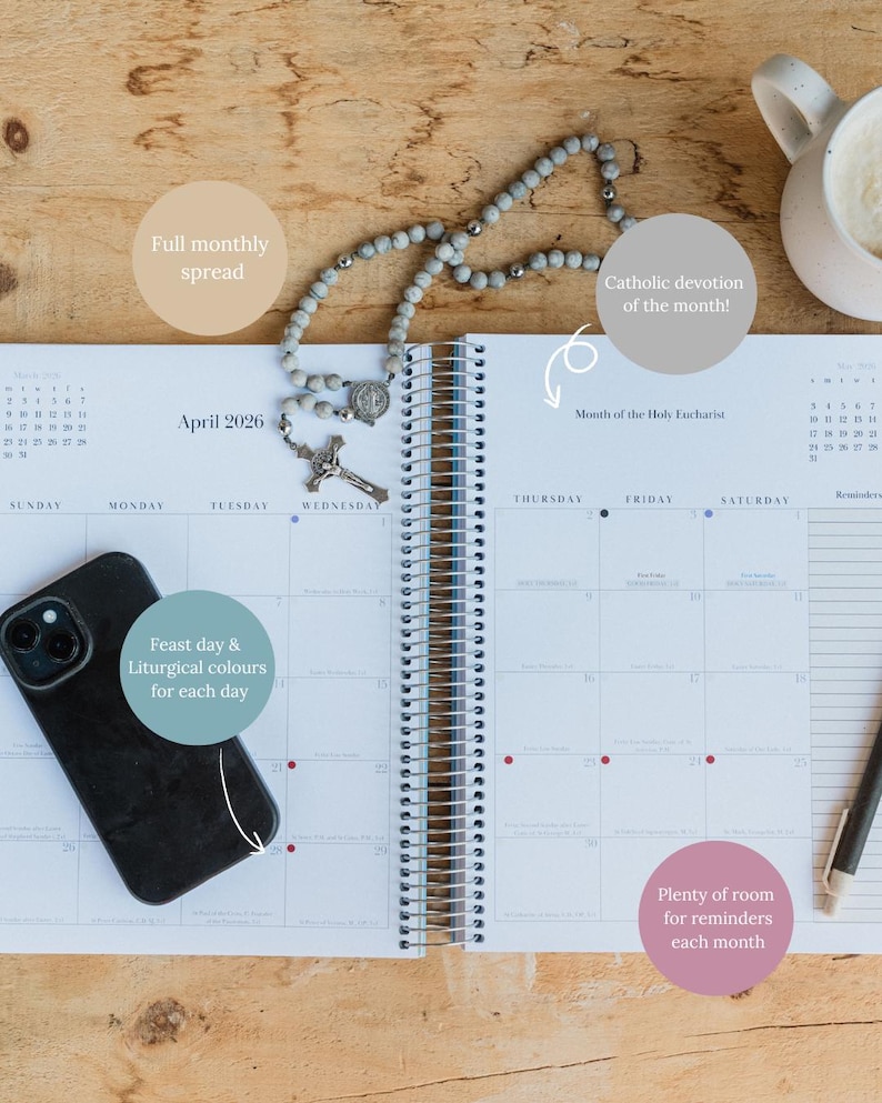 May include: Open spiral-bound planner for April 2026, featuring a full monthly spread, feast day and liturgical colors, and room for reminders. A rosary and a smartphone rest on the pages, with a mug of coffee nearby.