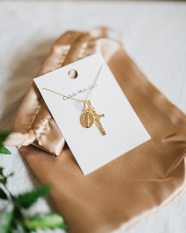 May include: Gold-colored necklace with a pendant featuring a cross and a medallion with a religious figure. The necklace is displayed on a white card with the text "Catholic Made Goods" and rests on a gold satin pouch.