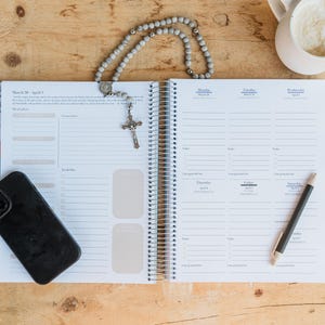May include: An open planner with a weekly layout, a black smartphone, a pen, a rosary, and a mug of coffee on a wooden surface. The planner is open to a week in March and April. The rosary has a silver crucifix.