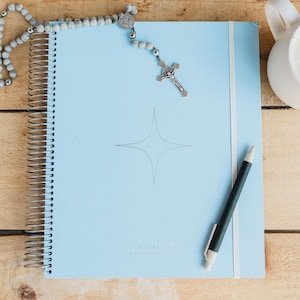 May include: A light blue spiral-bound calendar planner with a star design and the text "CALENDAR PLANNER". A black and white pen rests on the planner, along with a rosary and a white mug.