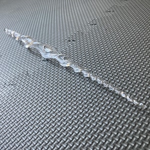 May include: A clear, transparent icicle-shaped ornament featuring a twisted, spiral design that tapers to a fine point at one end. The surface of the object is smooth and reflective, mimicking the appearance of frozen water in a long, slender form.