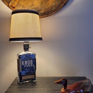 May include: A lamp made from a repurposed Knob Creek 12 bourbon whiskey bottle, featuring a beige lampshade with a dark trim. A wooden duck figurine sits on a dark wood surface. The lamp is on a dark wood table.