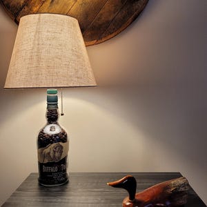 May include: A unique lamp made from a Buffalo Trace whiskey bottle filled with coffee beans, topped with a beige lampshade. A wooden duck figurine sits on a dark gray table next to the lamp. A wooden barrel head is mounted on the wall above.