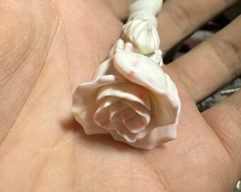 May include: A white carved rose with pink accents, the rose is attached to a long, thin stem.