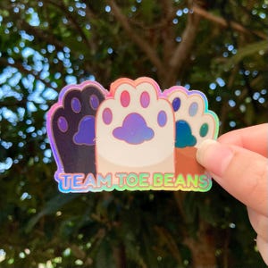 Peut inclure: Un autocollant holographique avec trois empreintes de pattes de différentes couleurs. Les empreintes de pattes sont disposées en rangée et l'autocollant indique "Team Toe Beans".