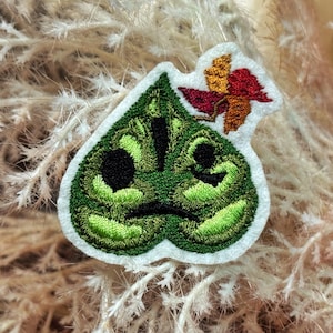 Op de afbeelding: Een groen geborduurde patch in de vorm van een blad met een gezicht, met zwarte ogen en een fronsende mond. Bovenop is een kleine, kleurrijke bloem bevestigd. De patch staat op een witte achtergrond.