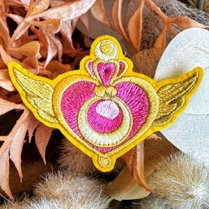 May include: Embroidered patch with a pink heart, gold crown and wings design, and a yellow border. The patch is set against a backdrop of dried flowers and foliage. The patch is likely for clothing or accessories.