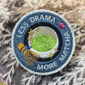 May include: Embroidered patch with a blue border and the text "LESS DRAMA MORE MATCHA". The patch features a green matcha bowl, a whisk, and a pink heart. The design is set against a background of white and brown foliage.
