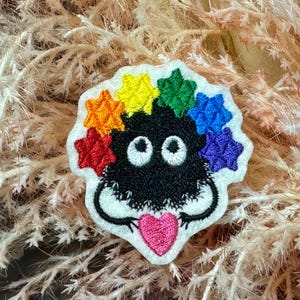 May include: Embroidered patch of a black creature with large white eyes and a pink heart-shaped tongue. It is surrounded by rainbow star-shaped accents. The patch is on a white background.