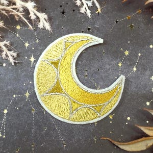 May include: Embroidered yellow crescent moon patch with starburst details, set against a dark grey background with constellation and star accents. The patch has a white border and is surrounded by dried foliage.
