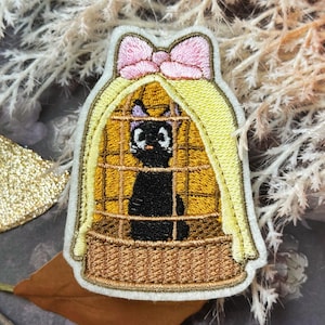 May include: Embroidered patch featuring a black cat inside a cage. The cage is yellow with a brown basket base and a pink bow on top. The cat has a white face and is looking forward. The patch is surrounded by a white border.