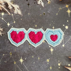 Può includere: Tre toppe ricamate a forma di cuore. Ogni toppa presenta un cuore rosso con un bordo azzurro. Le toppe sono disposte orizzontalmente, con i cuori che diminuiscono di dimensione da sinistra a destra. Lo sfondo è grigio scuro con accenti di stelle dorate.