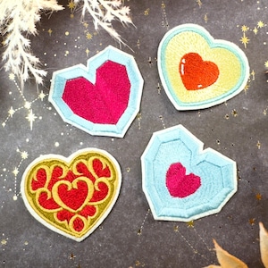 Puede incluir: Cuatro parches bordados en forma de corazón. Dos tienen un corazón rojo dentro de un borde geométrico azul claro. Uno tiene un corazón rojo con un borde amarillo. El último tiene un corazón rojo con un borde dorado.