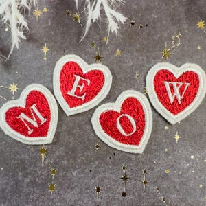 May include: Four heart-shaped embroidered patches spelling "MEOW". Each patch has a red heart with a white border and a white letter. The patches are arranged on a grey background with gold star accents and white floral elements.