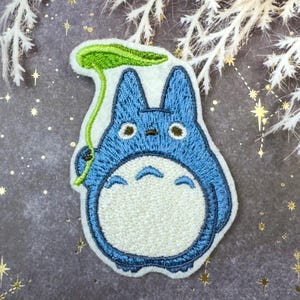 May include: Embroidered patch of a blue cartoon character with a white belly, holding a green leaf. The patch has a white border and is set against a grey background with star accents.