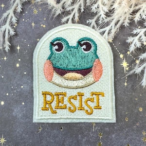 Puede incluir: Parche bordado con una rana sonriente con detalles en verde azulado y rosa. El parche tiene la palabra "RESIST" en letras doradas. El parche está sobre un fondo crema claro, con un elemento floral decorativo en el fondo.