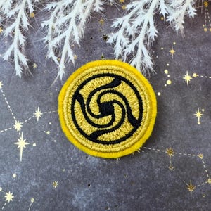 May include: A circular, yellow embroidered patch featuring a black spiral design. The patch has a black border. The background is dark grey with gold star constellations and white decorative branches.
