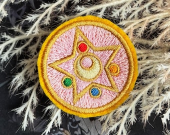 May include: Embroidered patch featuring a pink circle with a gold star and crescent moon design. The star has red, green, blue, and orange accents. The patch has a yellow border and is likely for clothing or accessories.