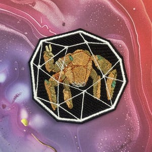 May include: Embroidered patch with a geometric web design, featuring a stylized insect. The insect is tan with green accents, set against a black background. The patch has a white border and is displayed on a colourful, marbled surface.