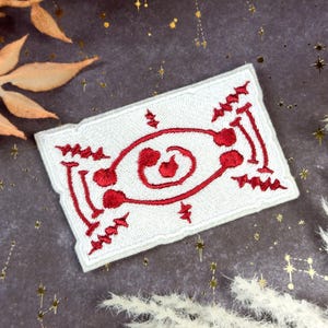 May include: White rectangular embroidered patch with a red abstract design. The patch features a central spiral and various linear and dot patterns. The edges are finished with a white border. The patch is likely for clothing or accessories.