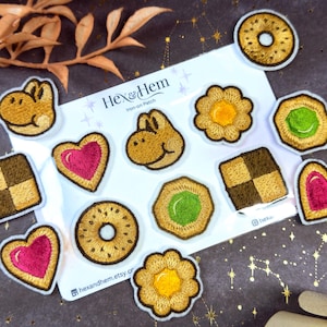 May include: A collection of embroidered iron-on patches in various shapes, including cookies, hearts, and a cartoon animal head. The patches feature detailed stitching in shades of brown, yellow, pink, and green, with a white border. The text "Hex & Hem" is visible.
