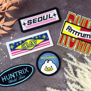 Puede incluir: Una colección de parches bordados con varios diseños y texto. Uno de los parches dice "SEOUL+" en rosa y blanco sobre un fondo negro. Otro dice "SLAY" con estrellas y rayas. Otros parches dicen "Rrrrrrumi", "HUNTRIX World Tour" y "Hello Friend" con un pato.