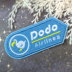 Puede incluir: Un parche bordado azul claro con las palabras "Dodo Airlines" en blanco y amarillo. El parche presenta un pájaro dodo de dibujos animados dentro de un círculo blanco. El parche es rectangular con esquinas redondeadas.