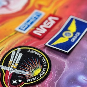 Może przedstawiać: Kolekcja haftowanych naszywek o tematyce kosmicznej. Zawiera naszywki z napisami "NASA", "GRACE" i "PROJECT HAIL MARY". Naszywki charakteryzują się żywymi kolorami i szczegółowymi obrazami, idealne do ozdabiania kurtek, toreb lub innych przedmiotów.