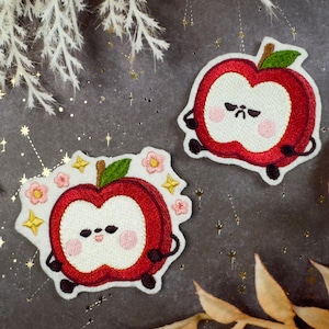 Pode incluir: Dois patches bordados de maçã, vermelhos e brancos. Um tem uma cara mal-humorada, o outro uma cara sorridente rodeada de flores e estrelas. Ambos têm braços e pernas pretos e folhas verdes.