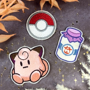 Op de afbeelding: Geborduurde patches met een rood-witte Poké Ball, een roze stripfiguur en een pot melk met een paarse deksel. De patches liggen op een donkergrijze achtergrond met gouden steraccenten en gedroogde bladeren.