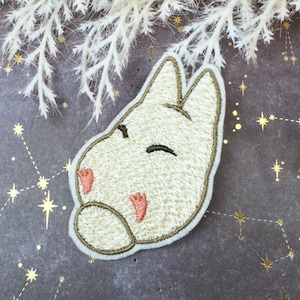 May include: Embroidered patch featuring a cream-colored bunny face with closed eyes and pink accents. The patch has a gold border and is set against a dark gray background with gold star constellations.