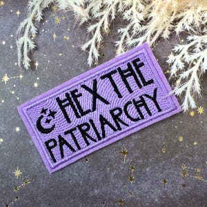 Op de afbeelding: Een rechthoekige, lavendelkleurige patch met zwarte geborduurde tekst "HEX THE PATRIARCHY". De patch heeft een maan- en sterontwerp. De achtergrond is een getextureerd grijs oppervlak met gouden steraccenten en witte bloemelementen.