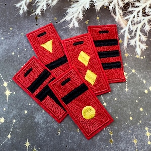 May include: A collection of red rectangular embroidered patches with black and yellow geometric designs. The patches feature various shapes, including triangles, diamonds, circles, and horizontal stripes. The patches are arranged on a textured, dark gray surface.