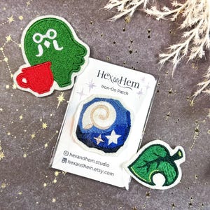 Puede incluir: Parches bordados para planchar. Uno presenta una silueta verde y una taza roja, otro un diseño azul y blanco con estrellas y una hoja verde. Los parches están sobre una superficie gris con un patrón de estrellas.