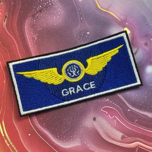May include: A blue embroidered patch with a white border. The patch features yellow wings, a circular emblem, and the name "GRACE" in white letters. The patch is rectangular in shape.