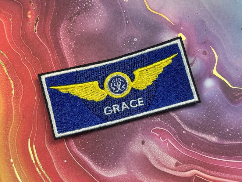 May include: A blue embroidered patch with a white border. The patch features yellow wings, a circular emblem, and the name "GRACE" in white letters. The patch is rectangular in shape.