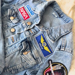 May include: Light blue denim jacket with multiple embroidered patches. Patches include the NASA logo, a blue and yellow winged emblem, and a patch that says "PROJECT HAIL MARY". The jacket has button closures.