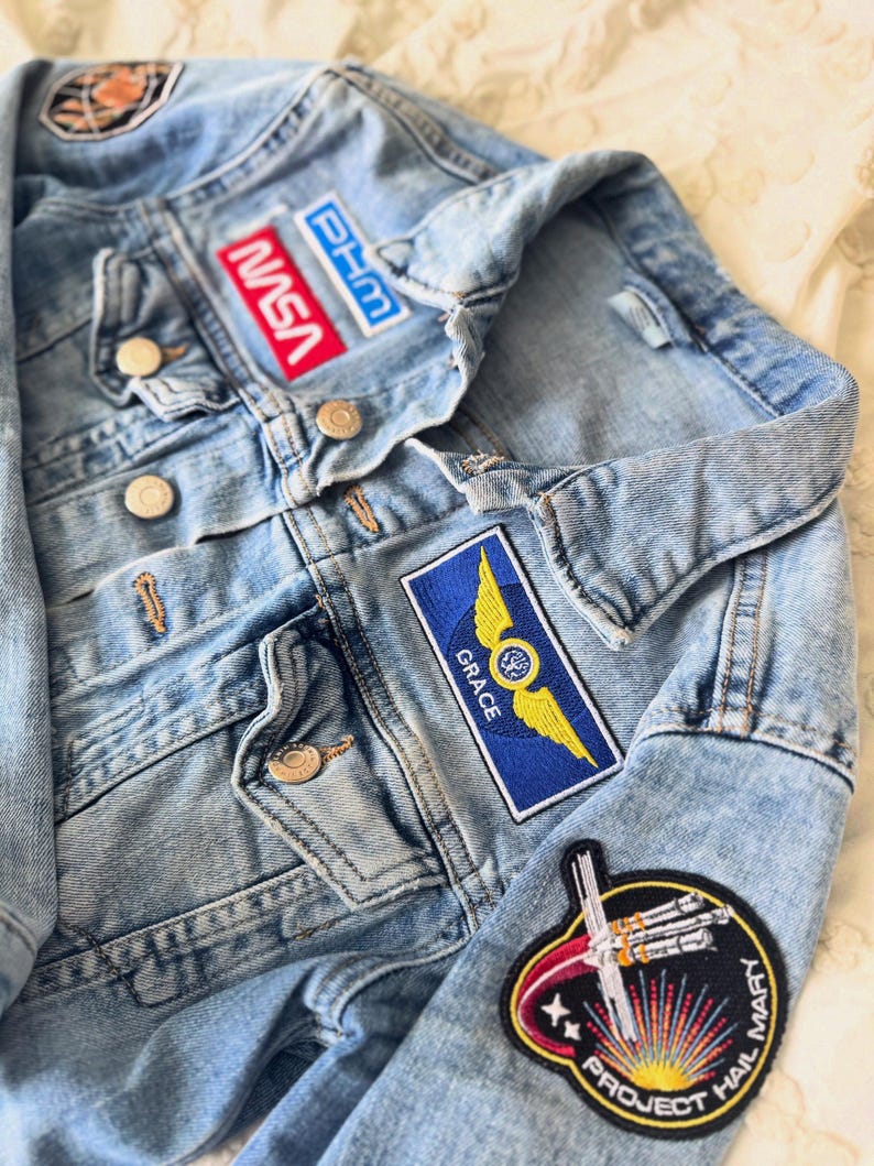 May include: Light blue denim jacket with multiple embroidered patches. Patches include the NASA logo, a blue and yellow winged emblem, and a patch that says "PROJECT HAIL MARY". The jacket has button closures.