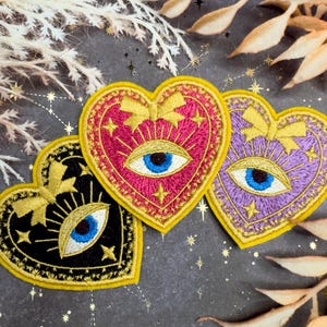 May include: Embroidered heart-shaped patches in black, pink, and purple, each featuring a blue eye design. Gold accents include stars, bows, and a yellow border. Decorative patches for clothing or accessories.