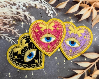 Puede incluir: Parches bordados en forma de corazón en negro, rosa y morado, cada uno con un diseño de ojo azul. Los detalles dorados incluyen estrellas, lazos y un borde amarillo. Parches decorativos para ropa o accesorios.
