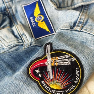 May include: Close-up of a light blue denim jacket with two embroidered patches. One patch is blue with yellow wings and the word "GRACE". The other patch is a circular design with the text "PROJECT HAIL MARY".
