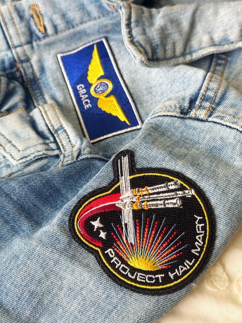 May include: Close-up of a light blue denim jacket with two embroidered patches. One patch is blue with yellow wings and the word "GRACE". The other patch is a circular design with the text "PROJECT HAIL MARY".