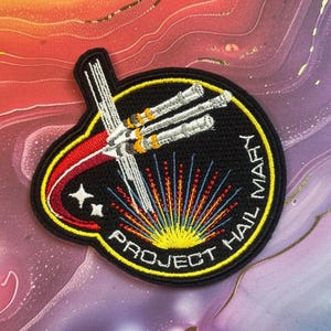 May include: Embroidered patch with a black background featuring a white spaceship design, red and yellow accents, and the text "PROJECT HAIL MARY." The patch has a yellow border and two white stars.