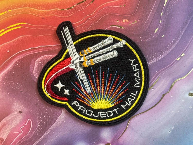 May include: Embroidered patch with a black background featuring a white spaceship design, red and yellow accents, and the text "PROJECT HAIL MARY." The patch has a yellow border and two white stars.