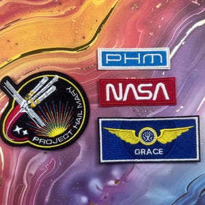 May include: A collection of embroidered patches with space-themed designs. Includes a patch with the text "PROJECT HAIL MARY", a NASA patch, a blue patch with gold wings and the name "GRACE", and a blue and white patch with the letters "PHM".