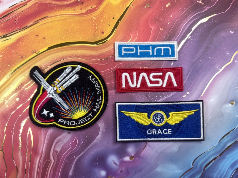 May include: A collection of embroidered patches with space-themed designs. Includes a patch with the text "PROJECT HAIL MARY", a NASA patch, a blue patch with gold wings and the name "GRACE", and a blue and white patch with the letters "PHM".