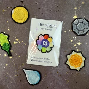 Puede incluir: Un surtido de parches termoadhesivos, que incluyen un c&iacute;rculo amarillo, una pluma verde, una flor multicolor, una forma geom&eacute;trica gris, un sol amarillo y una l&aacute;grima azul. Los parches est&aacute;n sobre un fondo oscuro con patrones de estrellas doradas.